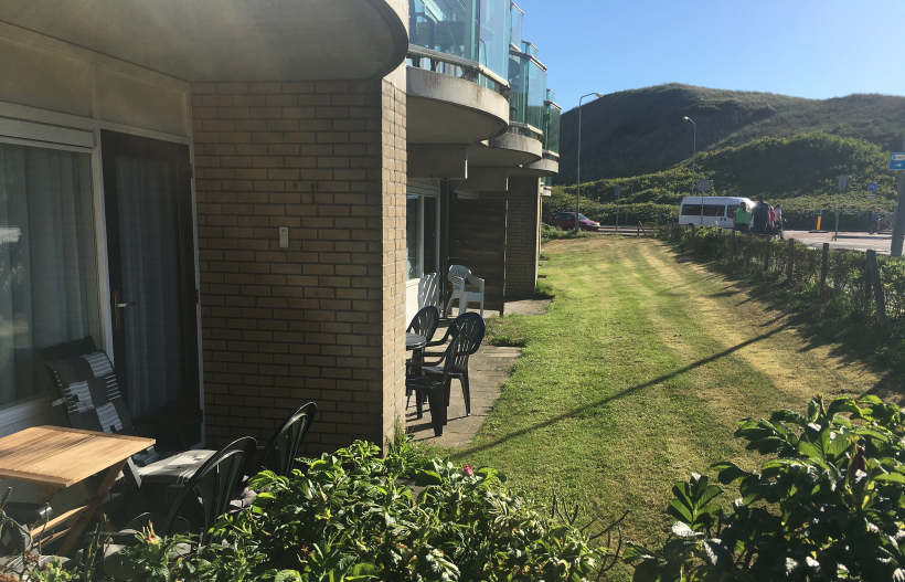 Foto Terrasse des Beach Appartements links im Bild, davor Wiese und im Hintergrund die Dünen, darüber blauer Himmel