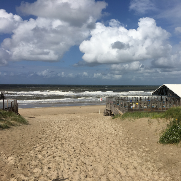 Foto Strandpavillion rechts im Bild, vorne Weg, dahinter Strand und Meer, darüber blauer Himmel mit weißen Wolken