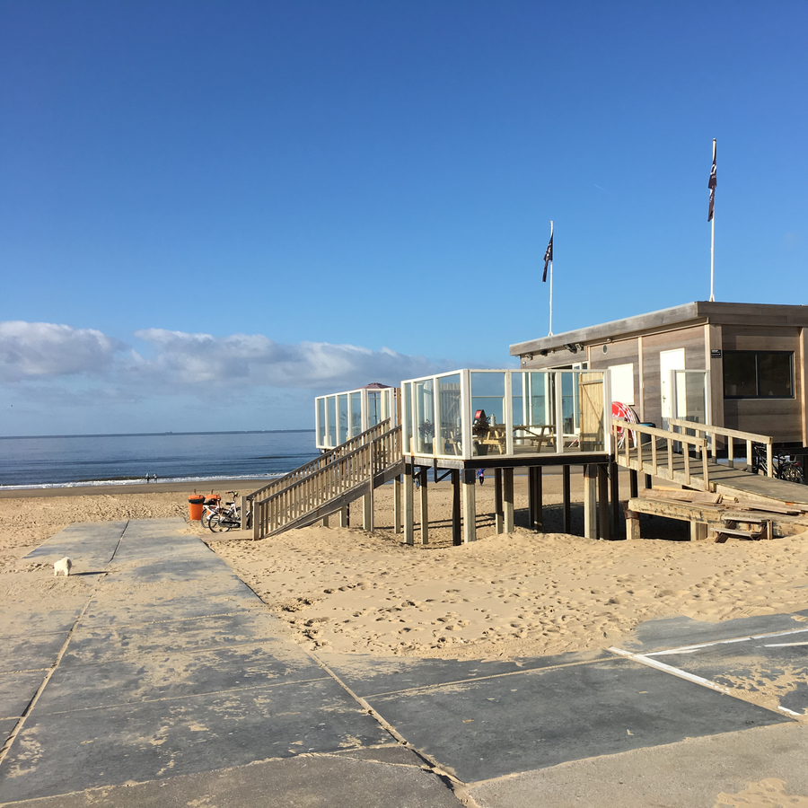 Foto Strandpavillion rechts im Bild, vorne Weg, dahinter Strand und Meer, darüber blauer Himmel